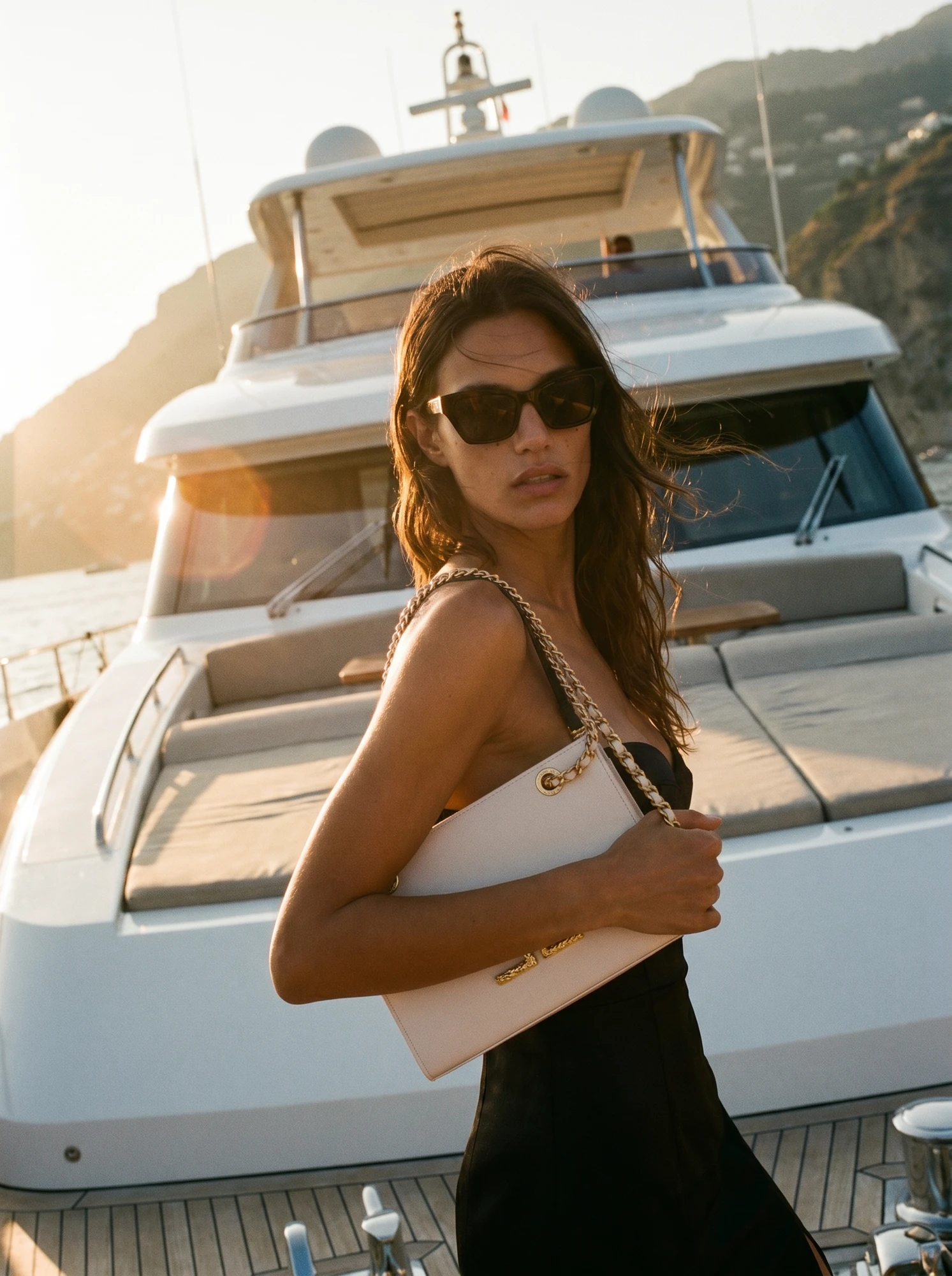 Modella con abito bianco e occhiali da sole davanti a uno yacht.